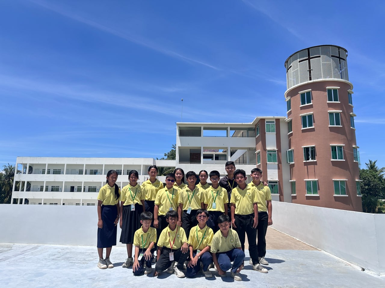 Youth Brigade students in yellow school shirts on the rooftop, with the classroom block and the round tower behind them
