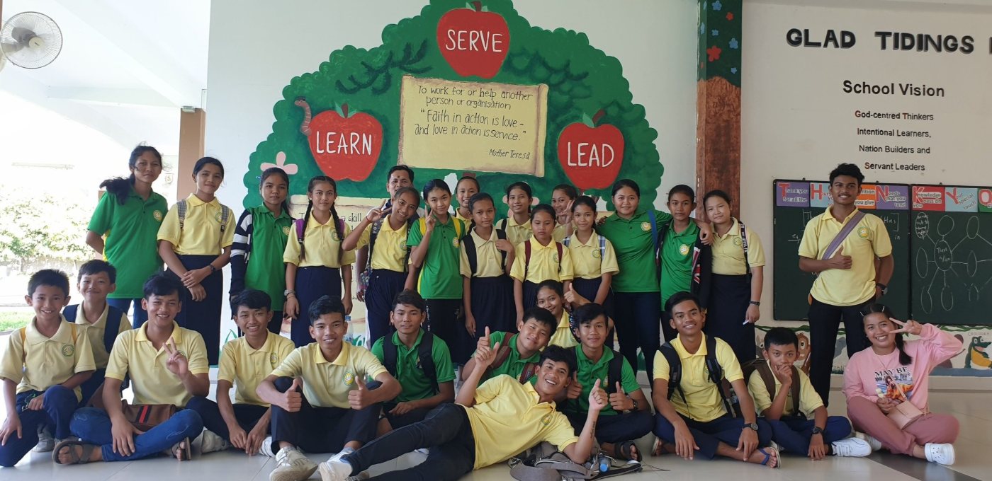 A class of GTIS pupils with their teachers in front of the painted school mural reading Learn, Serve, Lead, beside a board setting out the school vision